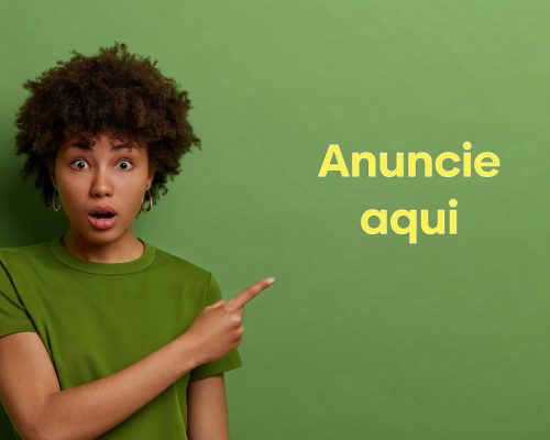 Surprised excited Afro American woman points index finger aside with shock, promots new product, looks with widely opened mouth at advertisement, wears bright green t shirt in one tone with background